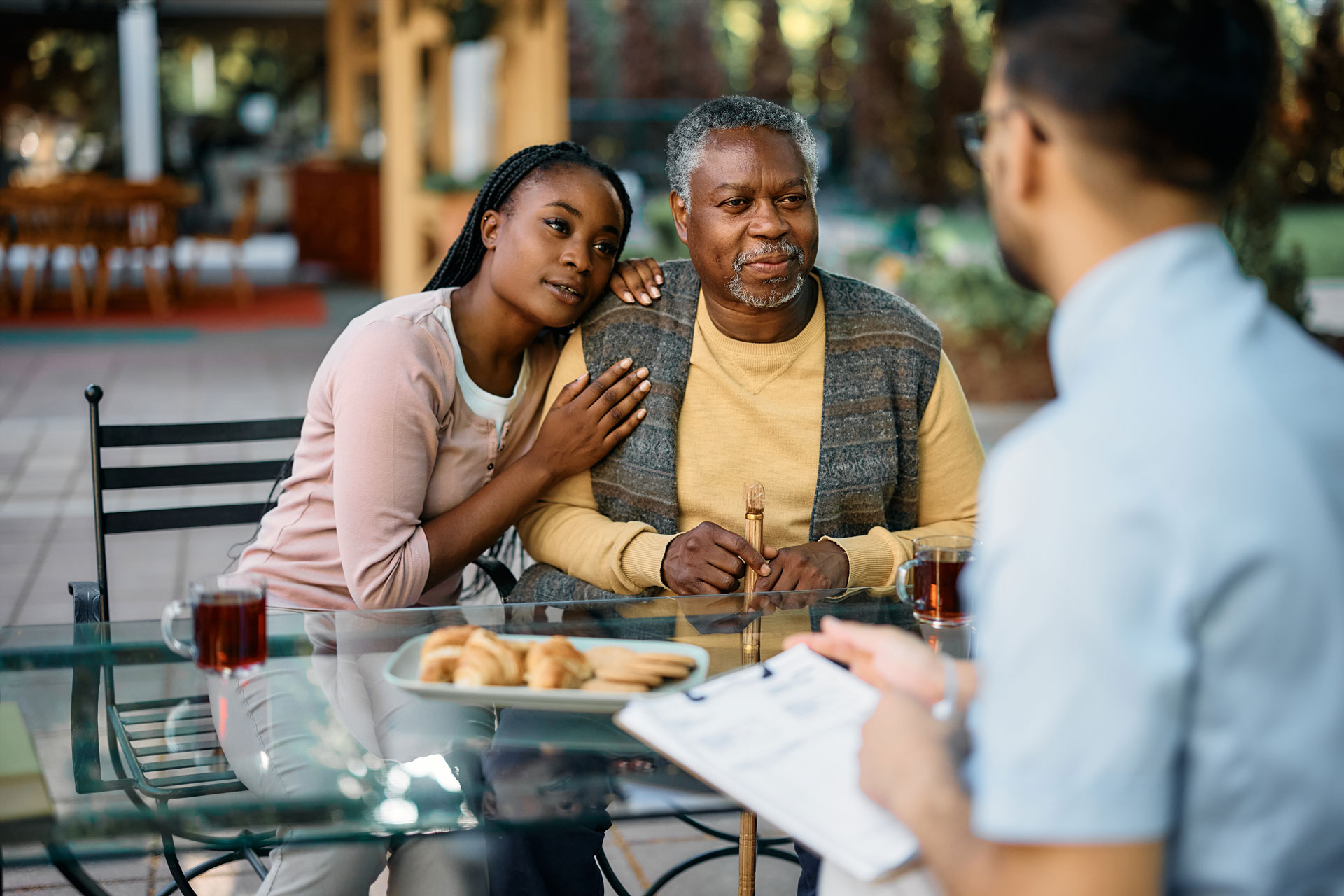 A senior man sitting at an outdoor table with his granddaughter leaning his shoulder.